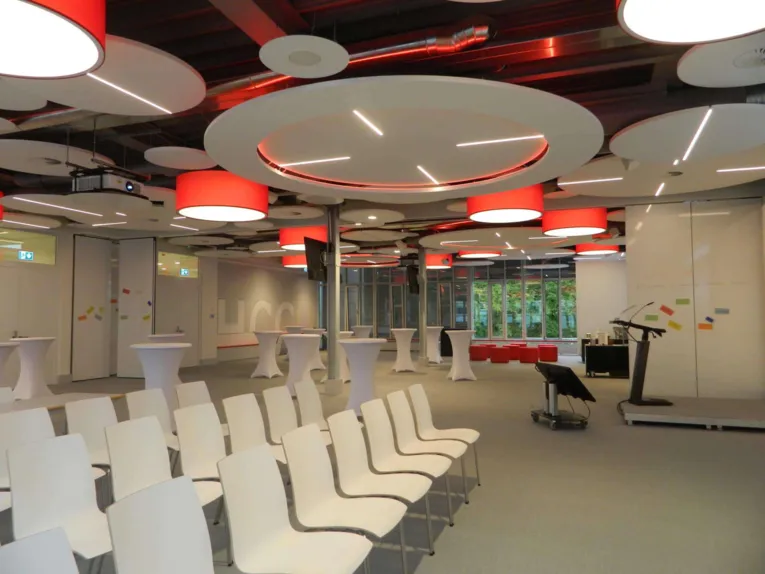 Moderner Konferenzraum mit weißen Stühlen, Stehtischen, Rednerpult und kreativer Deckenbeleuchtung in Rot und Weiß.